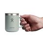 A hand holds a light green Hydro Flask Stainless Steel Mug (12 oz) with a splash-resistant lid against a white backdrop, featuring the brands logo of a person with raised arms.