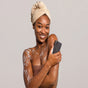 A smiling woman with a towel wrapped around her head holds a SheaMoisture African Black Soap Bar Soap with Shea Butter, washing her shoulder in rich lather from this natural skin cleanser against a plain background.