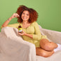 A smiling woman with curly hair sits on a beige chair against a green background, wearing a light yellow outfit and white socks. She holds a bottle of MegaFood Magnesium 300 mg and prepares to take one tablet.