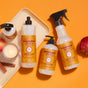 Mrs. Meyers Autumn Breeze Cleaning Set is displayed on an orange background with apples, cinnamon sticks, and a glowing candle, evoking a warm autumn vibe.