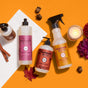 Four Mrs. Meyer’s cleaning products, a Soy Glass Jar Candle, pinecones, cinnamon sticks, red flowers, and autumn leaves are arranged on a marble slab with an orange background for a cozy fall-themed display.