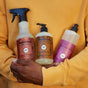 A person in a yellow sweatshirt holds three Mrs. Meyers products, including the Multi-Surface Cleaner, hand soap, and dish soap with colorful labels and plant-derived ingredients, close to their chest.