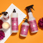Four Mrs. Meyer’s Autumn Breeze Cleaning Set products are arranged on an orange background with purple flowers and a lit candle for decoration.