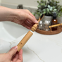 A person holds the Me Mother Earth All-in-One Bamboo Travel Toothbrush 2.0 Head - Refill with its matching case above a bathroom sink, alongside toiletries and a green plant in the background.
