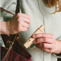 A person in a light green top holds a Me Mother Earth All-in-One Bamboo Travel Toothbrush 2.0 Head - Refill and a small bottle, while reaching into a maroon handbag with a brown strap.