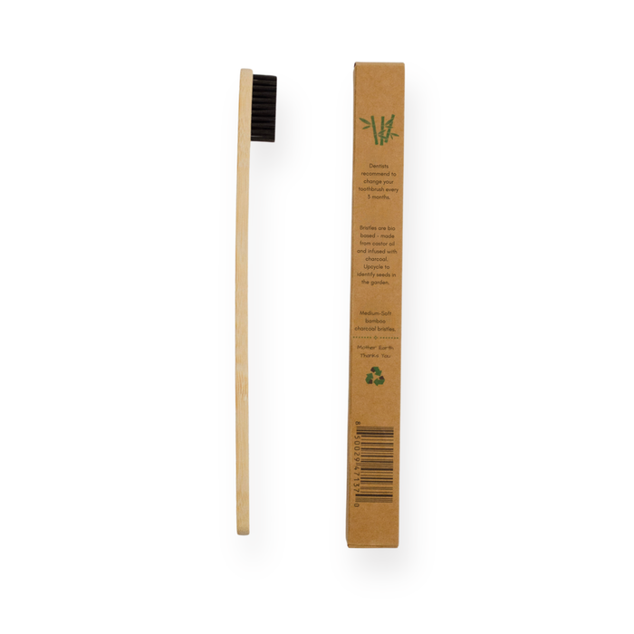 The Me Mother Earth Bamboo Charcoal Toothbrush with Castor Oil Bristles stands vertically beside its eco-friendly brown cardboard box on a white background—an eco-conscious choice for oral care.