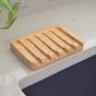 The Me Mother Earth Bamboo Draining Soap Dish with parallel grooves sits on a light-colored countertop near a sink, with a green plant visible in the corner.