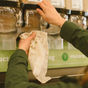 A person uses both hands to refill a Me Mother Earth Cotton Muslin Bulk Shopping Bag from a store dispenser. In the background, there's a green sign with partial text. The bag is part of the 3-Pack set.