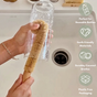 A person uses the Me Mother Earth Coconut Bottle Cleaning Brush with coconut husk bristles to clean a glass at the sink. Icons show it's reusable, eco-friendly, scrubby, and comes in plastic-free packaging.