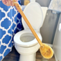A hand holds the Me Mother Earth Coconut Toilet Brush, made with eco-friendly coconut fiber and a sturdy wooden handle, above an open white toilet in a bathroom with a blue and white patterned shower curtain.