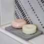 Two round solid bars, one pink and one white, sit on the Me Mother Earth Quick-Dry Diatomite Soap Dish labeled "Shampoo + Conditioner," beside a tiled wall with a decorative perforated metal panel.