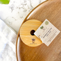 A Me Mother Earth Bamboo Mason Jar Lid with a logo rests on a wooden tray beside its tag, placed on white marble with a white cloth—ideal for anyone seeking eco-friendly jar lids.