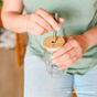 A person in a green shirt and jeans inserts a straw into a mason jar topped with a Me Mother Earth Bamboo Mason Jar Lid. Text in the image states: "Jar + Straw Not Included.