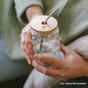 A person holds a clear mason jar fitted with a Me Mother Earth Bamboo Mason Jar Lid and metal straw, filled with water. Their hands rest on their lap. Text below reads: "*Jar + Straw Not Included.