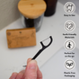 A hand holds a Me Mother Earth Charcoal Dental Floss Pick in a bathroom. Icons highlight its easy use, eco-friendly bamboo charcoal floss, ideal on-the-go design, and plastic-free packaging. A wooden jar and box are blurred behind.