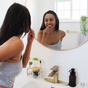A woman with long dark hair smiles as she brushes her teeth by a round mirror in a bright, modern bathroom. Me Mother Earth Charcoal Dental Floss Picks and a soap dispenser on the sink reflect her eco-friendly routine.