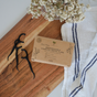 A box of Me Mother Earth Charcoal Dental Floss Picks rests on a wooden board beside eco-friendly floss picks, a white towel with blue stripes, and a small vase of flowers.