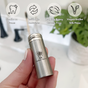 A hand holds a Me Mother Earth Refillable Charcoal Dental Floss container labeled Gentle on Gums, Refillable, Cruelty Free, Vegan Unlike Silk Floss. Ideal for eco-conscious users; bamboo charcoal floss inside. Blurred plant and sink in background.