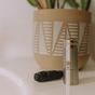 A Me Mother Earth Refillable Charcoal Dental Floss in a stainless steel container with logo sits on a white surface beside a black braided floss, with a large geometric-patterned plant pot in the background.