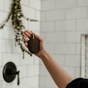 A hand holds up the Me Mother Earth Lava Pumice Stone—a natural foot scrubber with a white string—against white tiles, a black shower handle, and greenery. Upgrade your pedicure routine with this eco-friendly choice.
