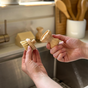 A person holds a Me Mother Earth Modular Sisal Multipurpose Brush handle and replacement head with plant-fiber bristles over a kitchen sink, with a bar of soap and utensils in the background.