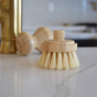 The Me Mother Earth Modular Sisal Multipurpose Brush Refill Head, made with plant-fiber bristles, is standing upright on a white countertop, with a wooden handle and brass faucet blurred in the background.