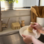A person cleans a white plate at the sink using Me Mother Earth’s Modular Sisal Multipurpose Brush, which features plant-based bristles. Nearby: a soap bar, dish rack, and utensil holder—all in sunlight for zero waste washing.