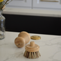 The Me Mother Earth Scrub Brush for Pots and Pans - Refill Head Only with natural bristles rests on a white marble countertop, with a glass vase of yellow flowers and a kitchen sink in the background.