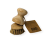 Two stacked Me Mother Earth Scrub Brushes for Pots and Pans with plant-based bristles are tied with string and attached to a brown product tag featuring text and a barcode.