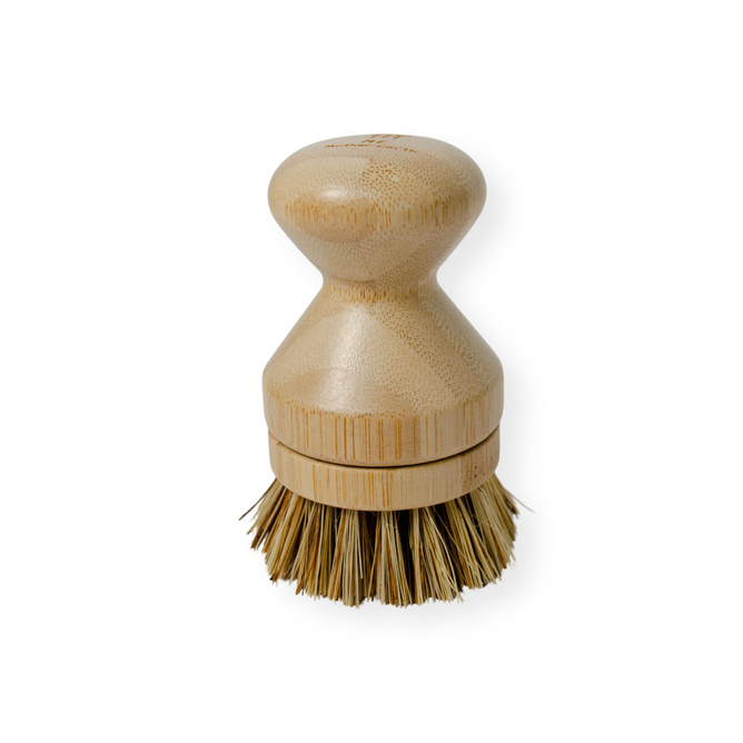 The Me Mother Earth Scrub Brush for Pots and Pans features a short, smooth bamboo handle and stiff plant-based bristles at the bottom for effective cleaning. This plastic-free brush is shown against a white background.