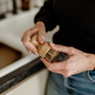 A person cleans the Me Mother Earth Scrub Brush for Pots and Pans—featuring a bamboo handle and plant-based bristles—with their fingers over a kitchen sink, while wearing blue jeans and a dark top.