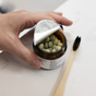 A hand holds an open amber jar of Me Mother Earth Mouthwash Tablets - Zero Waste, with green tablets next to a bamboo toothbrush on a white surface—a simple, plastic-free oral care solution.
