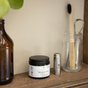 A jar of Me Mother Earth Mouthwash Tablets - Zero Waste, a metal tin, a glass jar with a bamboo toothbrush, and a brown bottle are arranged on a wooden shelf against a beige wall.