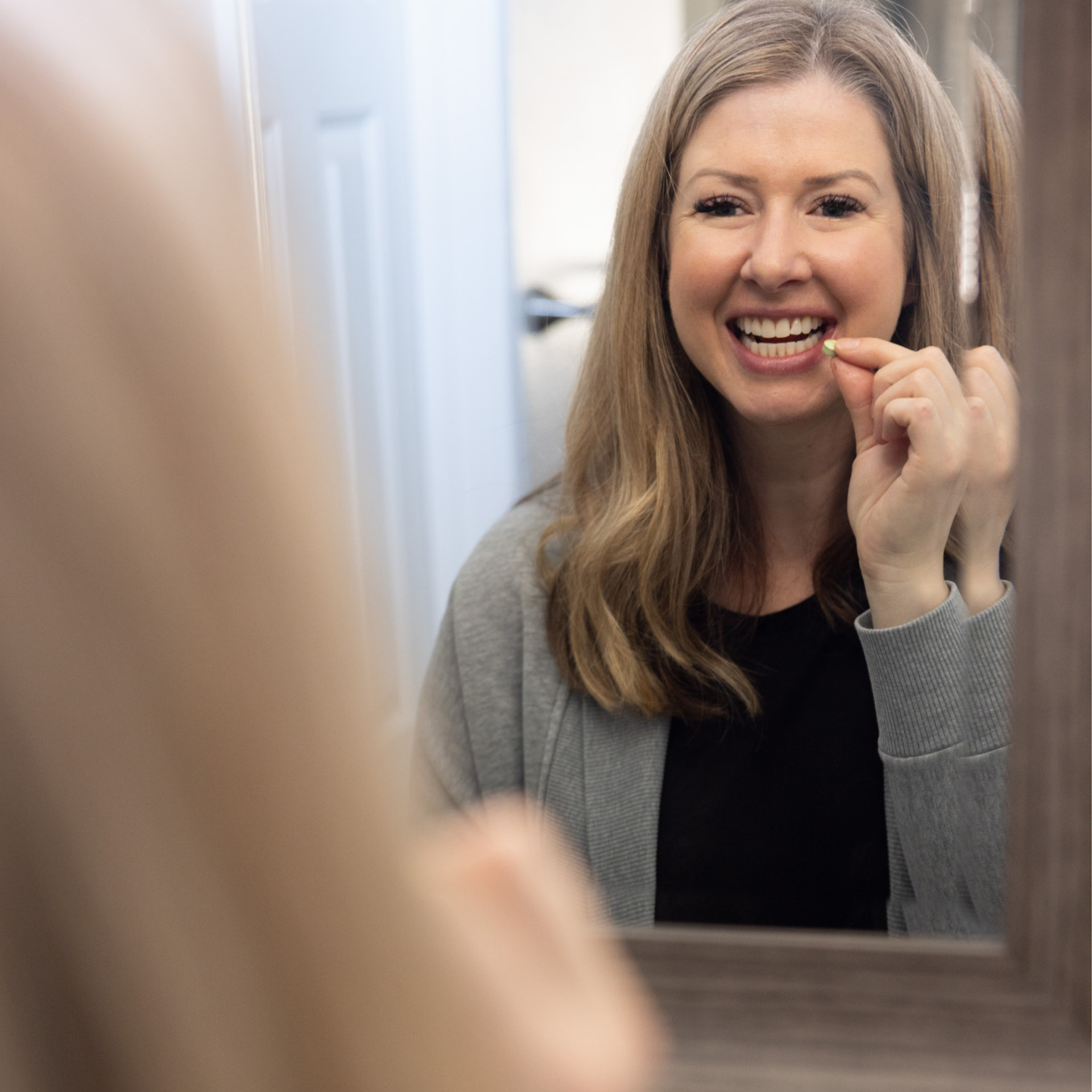 A woman with long blonde hair smiles in the mirror, using dental floss. On the counter are Me Mother Earth Mouthwash Tablets - Zero Waste, part of her fresh oral care routine. She wears a gray cardigan over a black shirt.