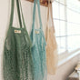 Three "One Tripper" Expandable Mesh Market Bags by Me Mother Earth—in green, blue, and beige—hang by a window. Made from organic cotton, each bag catches sunlight, adding a bright and airy feel to the space.