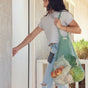 A woman in a striped shirt and ripped jeans opens a door while carrying the Me Mother Earth "One Tripper" Expandable Mesh Market Bag filled with groceries like apples and cabbage.