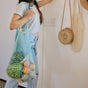 Wearing a striped crop top and ripped jeans, a person holds Me Mother Earth's "One Tripper" Expandable Mesh Market Bag filled with produce, standing indoors by a wall decorated with hanging woven bags.