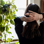 A person with long brown hair stands indoors by a leafy plant, gathering their hair with both hands to tie it back using Me Mother Earth Hair Ties made from cotton and natural rubber, as soft light filters through the window.