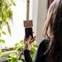 A person with long brown hair holds a pack of Me Mother Earth Hair Ties made from cotton and natural rubber near a window, surrounded by green potted plants.