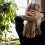A person with blond hair, seen from behind near a sunlit window and lush green plants, gathers their hair using Me Mother Earth Hair Ties made from cotton and natural rubber.