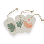 Three rectangular tags with rounded corners and string loops, each with minimal designs—a green monstera leaf, a cactus with pink flowers, and abstract pink-orange shapes—are perfect accents for Me Mother Earth's Pop Up Dish Sponges 3-Pack.