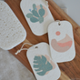 Three rectangular off-white tags with string loops, featuring minimalist plant or abstract pastel designs, lie on a wooden board next to a Me Mother Earth Pop Up Dish Sponges 3-Pack and a pumice stone.