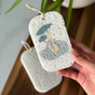 A hand holds a Me Mother Earth Pop Up Dish Sponge from the 3-Pack set, shown in textured white with blue and beige mushroom illustrations; another sponge is blurred in the background on a wooden surface.