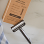 A matte black Me Mother Earth Reusable Double Edge Safety Razor sits next to its brown eco packaging, featuring zero waste shaving, a smooth shave, a cotton travel pouch, and reduced waste.