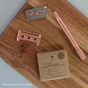 A brown box labeled "Me Mother Earth Safety Razor Refill Pack," rose gold razor head, and stainless steel replacement blades are displayed on a wooden board. Text reads: *Safety Razor Not Included.