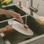 Someone washes a white plate under running water in a kitchen sink using the Me Mother Earth Sisal Kitchen Brush. Nearby, there’s a soap bar, another brush, and some vegetables.