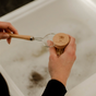 A person uses a beechwood-handled jar opener to open a glass jar labeled Me Mother Earth above a white sink filled with soapy water, next to Me Mother Earth's Sisal Kitchen Brush Refill Heads.