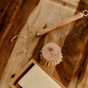 A Me Mother Earth sisal kitchen brush refill head rests alongside a beechwood dish brush, metal and wood weeding tool, bar of soap, and wooden soap dish on a wooden surface.