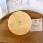 A Solid Bamboo Mason Jar Lid by Me Mother Earth with an engraved tree logo sits on a wooden surface beside a branded business card, highlighting eco-friendly bamboo mason jar lids. A white cloth and marble counter are also partially visible.