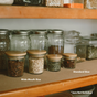 Solid Bamboo Mason Jar Lids by Me Mother Earth are shown on glass jars filled with dried foods on wooden shelves; available in Wide Mouth and Standard Size. Text in the corner states *Jars Not Included.